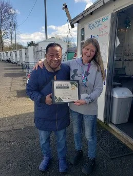 Two people, a man and a woman, smiling and holding a certificate outdoors in front of a white building with chalkboard signs.