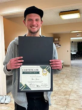 A young man smiling and holding a certificate in a black folder inside a building with tiled flooring and a ceiling light.