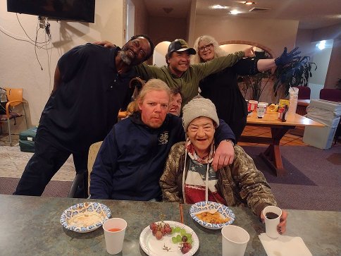 A group of five people gathered around a dining table in a casual indoor setting, smiling and posing for the photo. There are plates with food, cups, and a small cake with grapes on the table.