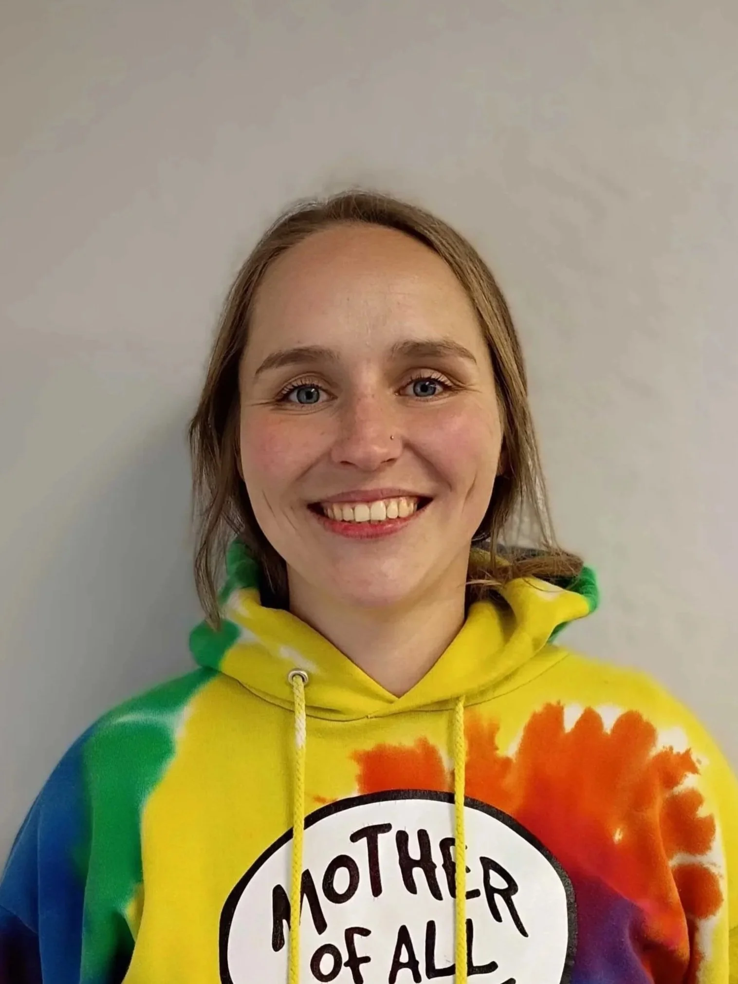 A smiling young woman with light skin, blue eyes, and shoulder-length light brown hair, wearing a colorful tie-dye hoodie with the words "Mother of All Things" on it, standing against a plain off-white wall.