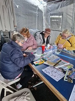 Four women sitting around a table inside a tent, looking at papers and working on a craft project, with various supplies and magazines on the table.