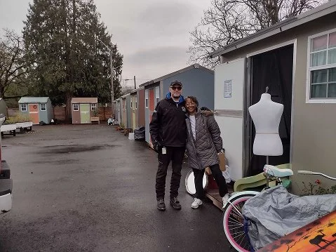 Two people standing outdoors in front of small buildings, with a mannequin and a bicycle nearby.