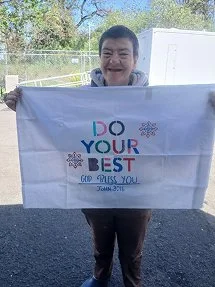 Person smiling and holding a white banner that reads 'Do Your Best, God Bless You,' outdoors with trees and a fence in the background.