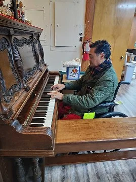 A man sitting and playing an upright piano in a room with wooden flooring and a door in the background.