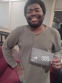 A smiling young man with a beard holding a black card with 'SOS' written on it, standing indoors near a window.