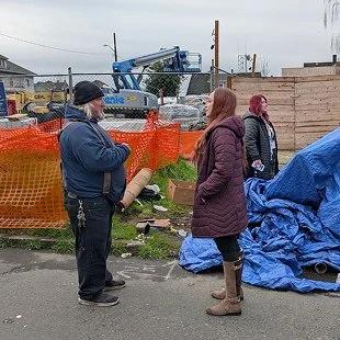 Police officer talking to two women near a construction site with orange fencing and blue tarps.