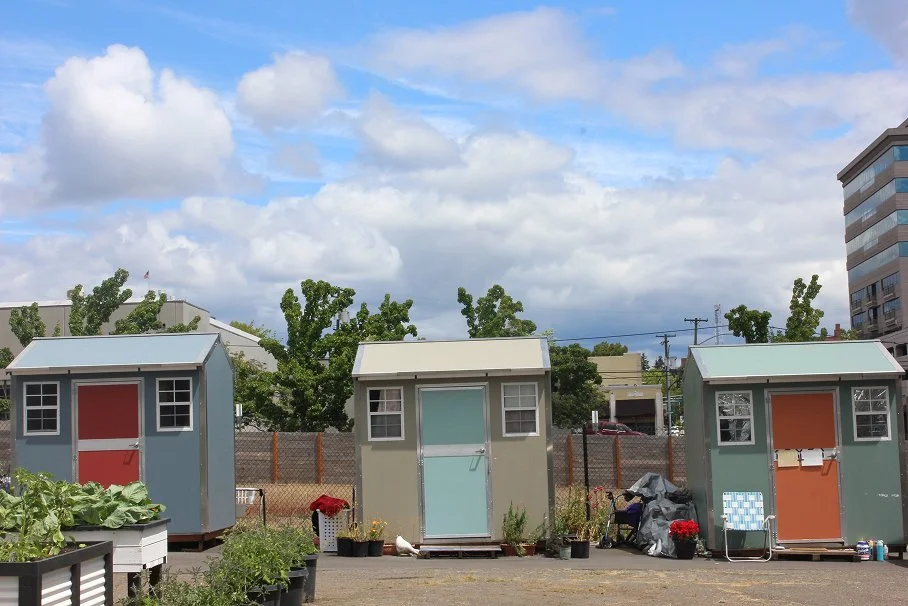 Three small colorful safe stay units arranged side by side outdoors, with greenery, potted plants, and a blue sky with clouds in the background.