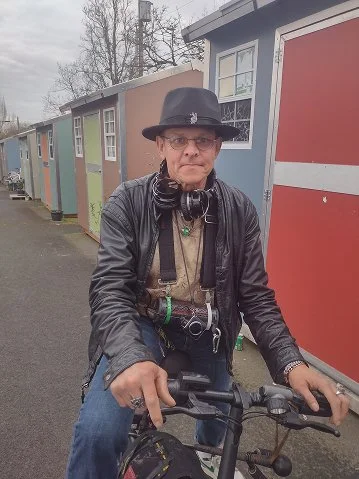 A person wearing a black hat, glasses, a leather jacket, and headphones around their neck, riding a bicycle on a street lined with colorful tiny houses.