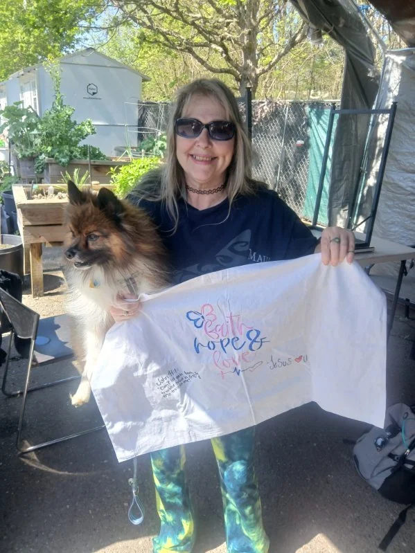 Woman with sunglasses holding a dog and a white cloth with handwritten messages outdoors on a sunny day.
