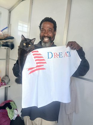 A man smiling and holding a white shirt with the word 'DREAM' and a stencil of the American flag on it, with a black cat sitting on his shoulder.