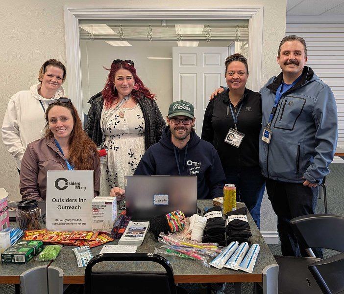 Group of six people standing around a table with outreach materials, smiling, in a room with a window and door in the background.
