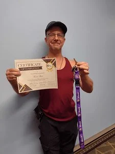 Smiling man holding a certificate and medal, wearing a cap and athletic clothing, standing in front of a plain wall.