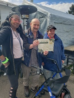 Three people standing outdoors, one holding a certificate, with a tent in the background.