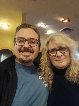 A man and woman taking a selfie indoors. Both are wearing glasses and smiling.