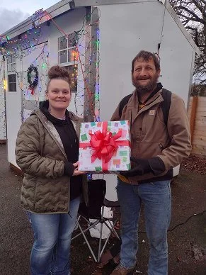 A young woman and a man holding a gift box with a red bow, standing outside decorated with Christmas lights and a wreath, during winter.