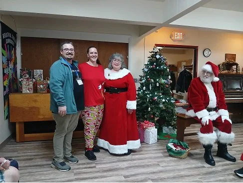 Three people standing together with a Christmas tree and Santa Claus in the background, in a festive indoor setting.