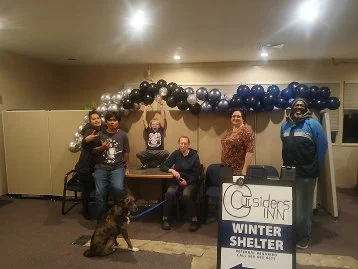 Group of five people standing and sitting behind a table at an indoor event, with a black and white sign that reads "C Sidder's Inn Winter Shelter" in front. There are black, gray, and blue balloons above them, and a dog sitting in front.