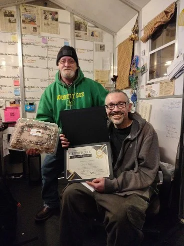 Two men inside a decorated room, one sitting holding a certificate and the other standing holding a large snack. The seated man has glasses, a beard, and gray hair, and the standing man is wearing a black beanie and green hoodie that says 'Country Bo
