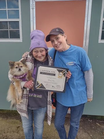 Two women, one holding a small dog, stand outside near a building, smiling at the camera. The woman on the left is wearing a purple beanie and holding a certificate, while the woman on the right is wearing a blue scrub top and cap.