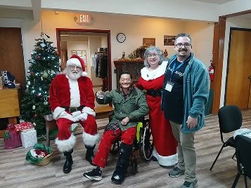 Four people, including Santa Claus, two women dressed as Santa, and a man, posing together indoors near a decorated Christmas tree with presents, during a holiday celebration.