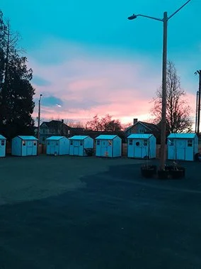 A row of small blue beach huts with white doors lined up along a grassy area during sunset, with trees, houses, and streetlights in the background.