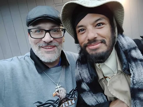 Two smiling men taking a selfie together. The man on the left has glasses, a beard, and is wearing a cap, a necklace with a cross, and a gray shirt. The man on the right is wearing a hat, a plaid jacket, and a light-colored shirt.