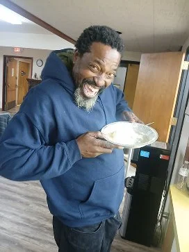 A man with curly hair and a beard smiling and holding a clear bowl of food in a kitchen.