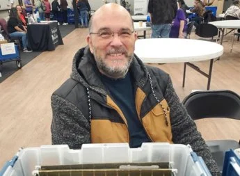 Man with glasses and beard smiling, sitting at a table in an indoor event space.