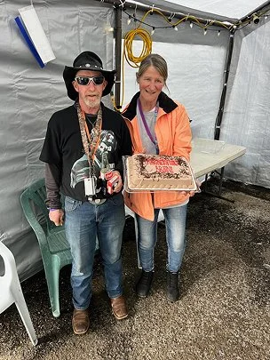 A man and woman standing inside a tent, holding a large cake, with the woman smiling and the man wearing sunglasses and a hat.
