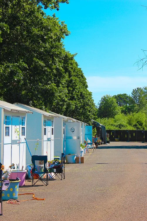 A row of blue beach huts with white doors, some with window curtains, along a paved area under a large green leafy tree, with people sitting on chairs and a black truck in the background under a bright blue sky.
