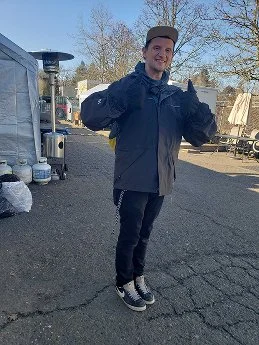 Young man standing outdoors on a paved area, smiling, wearing a dark jacket, black pants, sneakers, and giving two thumbs up.