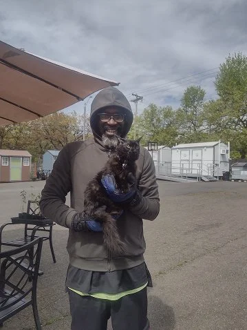 Man in hoodie holding a small black dog outdoors under a cloudy sky.