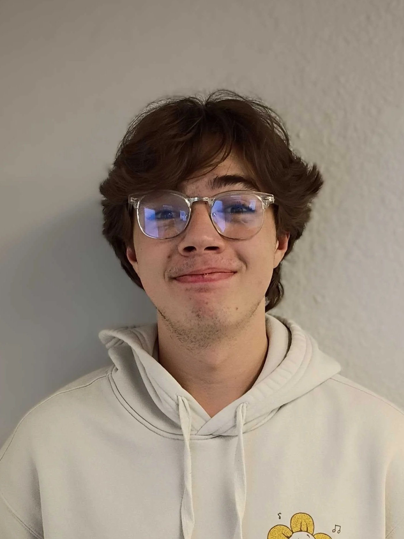 A young man with brown hair and glasses smiling while wearing a white hoodie with a flower cartoon design on the front. He stands against a light-colored textured wall.