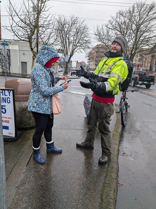 A man in a bright yellow rain jacket talks to a woman in a blue patterned coat on a wet city sidewalk. The woman is using her phone, and the man is gesturing with his gloved hands. There are bicycles, a black truck, leafless trees, and buildings in the background, indicating a cold, rainy day.