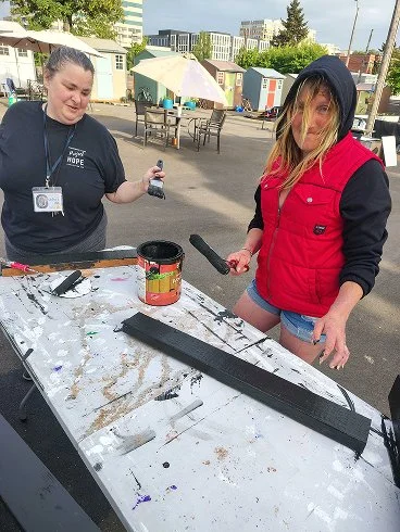 Two women working on a wood project outdoors. One woman is holding a hammer and the other woman is holding a tool, with a long piece of wood on a table covered with paint splatters. In the background, there are tables, umbrellas, and buildings.