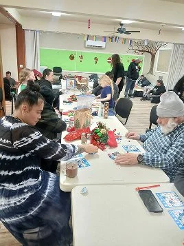 Group of people sitting around tables engaging in a craft activity, with Christmas decorations and supplies on the tables, in a decorated room.