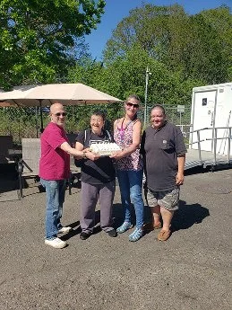 Four women standing outdoors around a birthday cake, smiling and holding the cake. Background includes a large umbrella, trees, a trailer, and a clear blue sky.