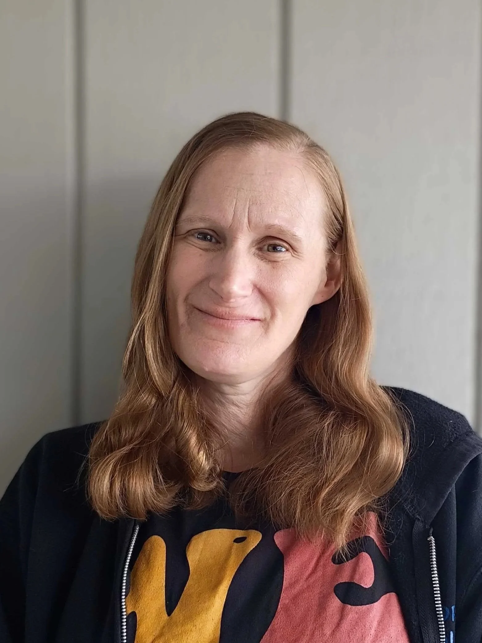 A woman with long, wavy red hair smiling against a plain, light-colored wall.