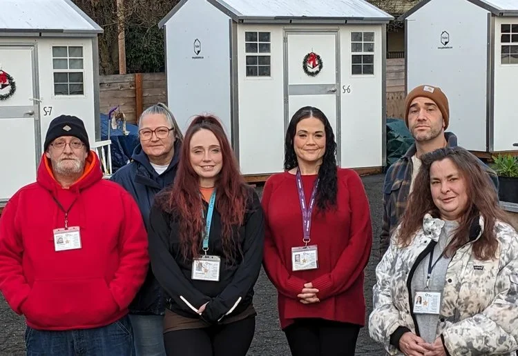 Six people standing outdoors in front of small white and gray shed structures decorated with holiday wreaths, wearing ID badges and winter clothing.
