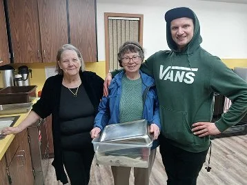 Three people standing together in a kitchen, smiling, with one holding a shiny tray or container.