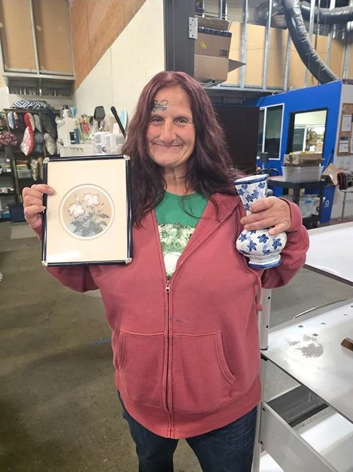 A woman with long dark hair, wearing a red hoodie and green shirt, smiling and holding a framed artwork and a ceramic mug in a store or warehouse setting.