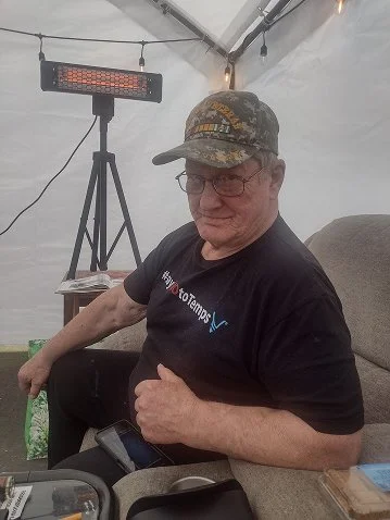 Man sitting on a chair under a tent, wearing a camouflage cap and black shirt, with a red LED heater or light behind him.