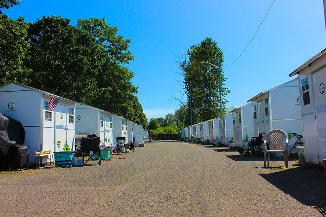 Row of white tiny houses or cabins along a gravel road with outdoor chairs, tables, and bicycles outside, under a clear blue sky with green trees in the background.
