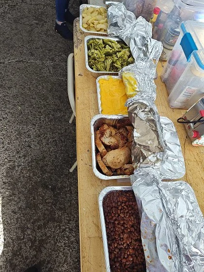 Aluminum trays on a wooden table filled with potatoes, broccoli, melted cheese, roasted chicken, and cooked beans, with foil partially covering some trays.