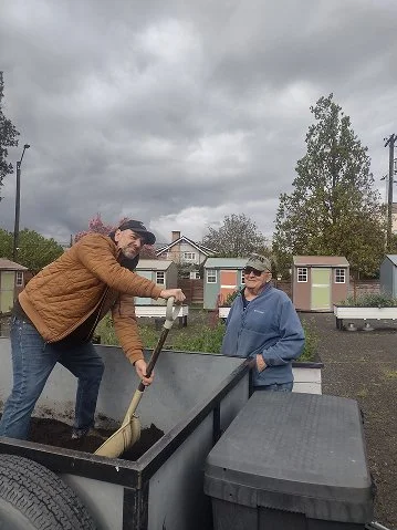 Two men working outdoors with a wheelbarrow and gardening tools on a cloudy day in a residential area.