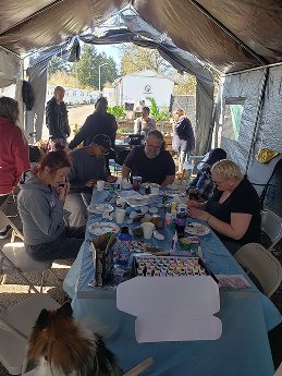 People sitting at a long table inside a large outdoor tent, engaged in arts and crafts activities with various supplies on the table.