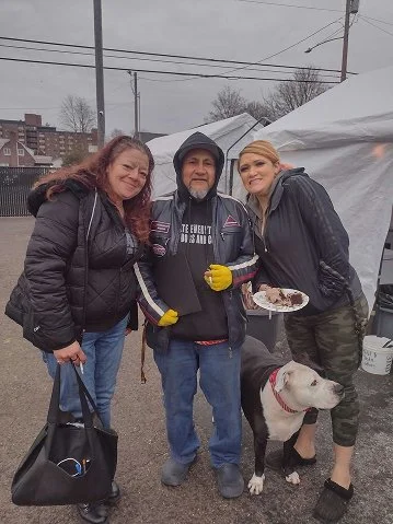 Three people, a dog, and a white tent outdoors on a cloudy day. The man in the middle is holding a plate of food and the dog is sitting on the ground. Two women are standing beside him, one holding a black bag.