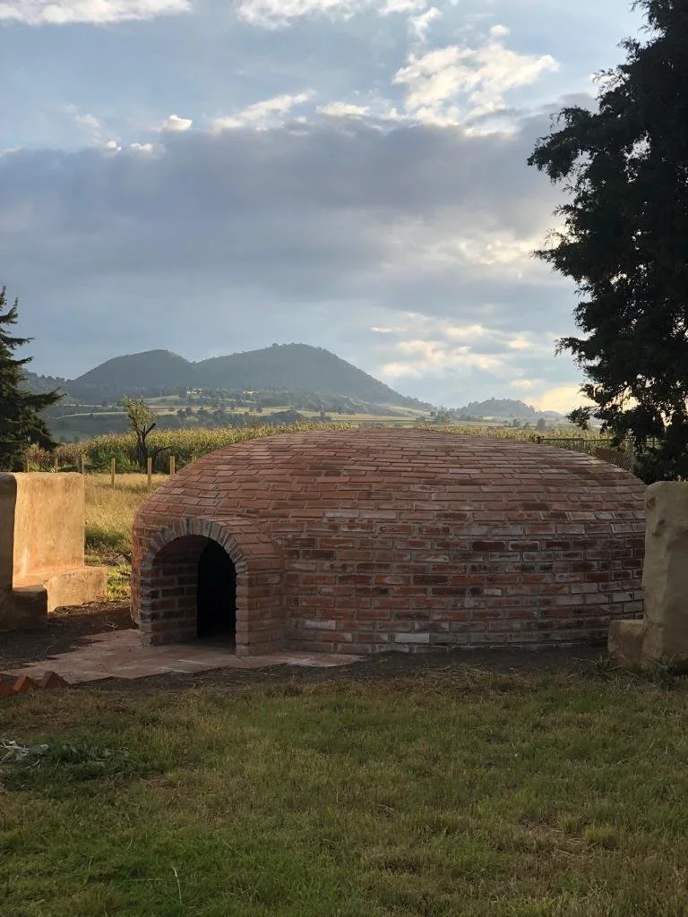 Una estructura de ladrillos en forma de horno o iglú en un área de césped con árboles y montañas de fondo bajo un cielo con nubes.