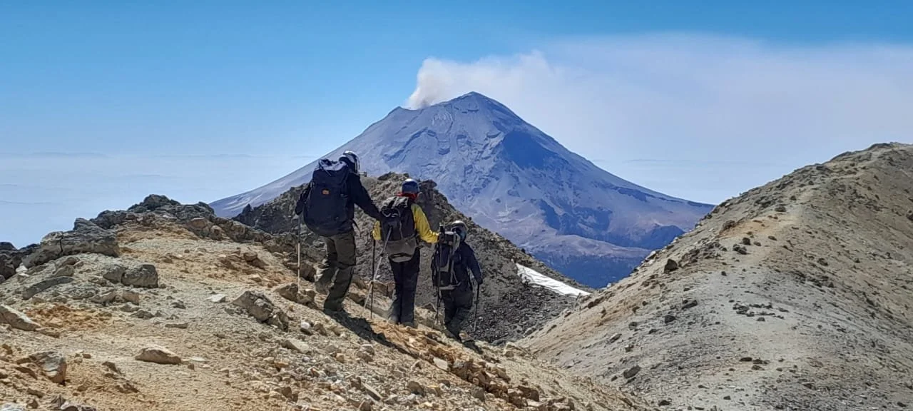 Tres montañistas avanzando por un sendero rocoso en un volcán activo con humo saliendo de su cráter, en un paisaje montañoso con cielo despejado.