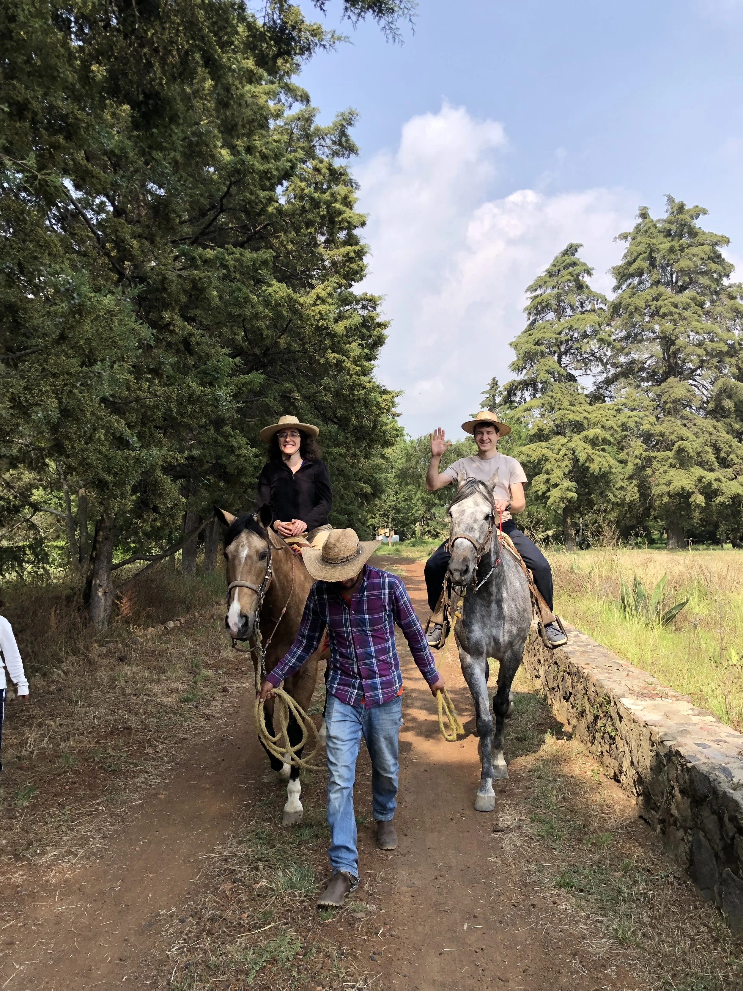 Dos personas montando caballos y una persona guiándolos por un camino en un entorno natural con árboles verdes y cielo nublado
